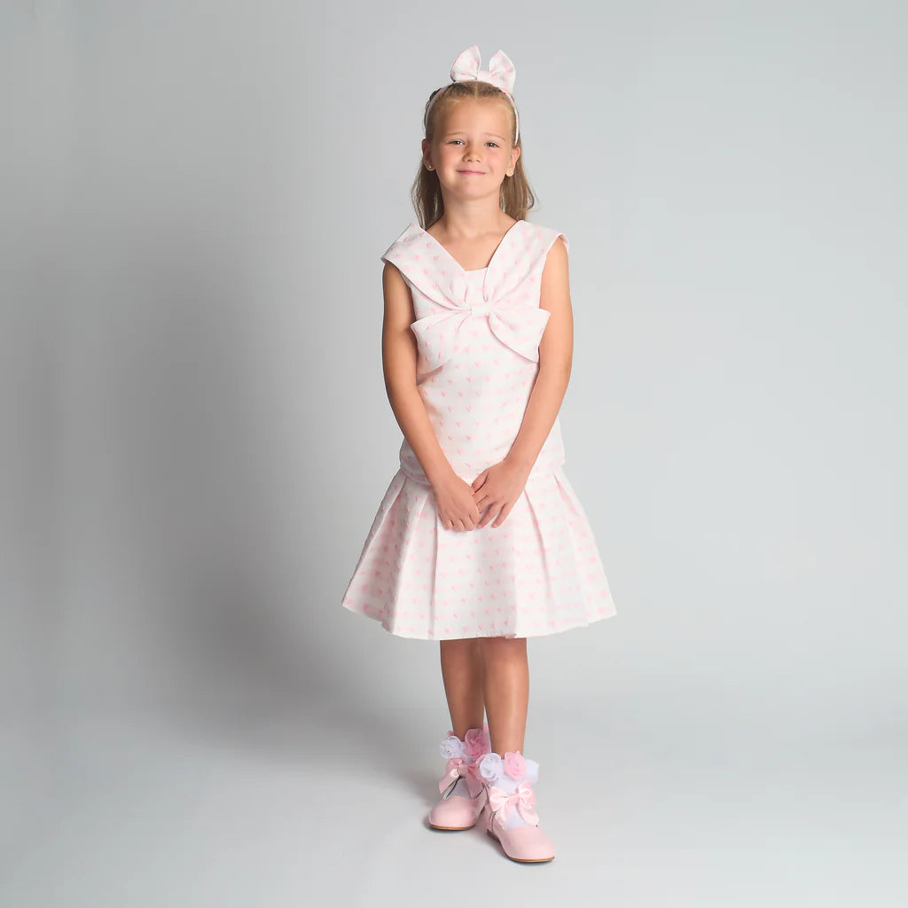 Full-length studio shot of the Beau Kid girls pink heart jacquard skirt set, showcasing the structured chest bow and box pleats.