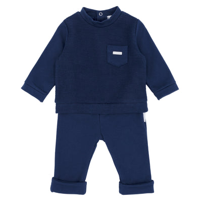 Blues Baby branded boys navy blue two piece jogger set finished in American fleece fabric. This set consists of a long sleeve top, which has a small pocket detail to the front and push button fastening on the rear. It comes matching trousers also. Available in sizes 3 month up to 24 month.  Also available in baby blue colour.