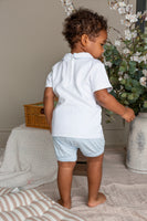 Child in a white outfit standing near a plant and basket in a room.
