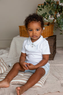 Child wearing a white shirt and light blue shorts sitting on a striped blanket with wicker baskets and plants in the background.