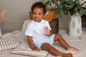 Child in a white shirt and diaper sitting on a bed with decorative pillows and plants in the background.