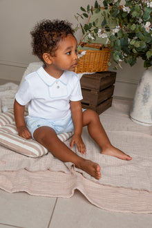 Child sitting on a bed wearing a white shirt and light blue shorts, with a plant and wooden basket in the background.